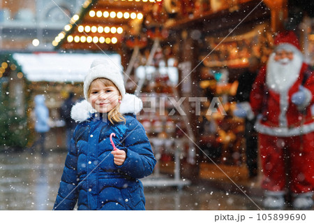 Little cute preschool girl with candy cane from a sweets stand on Christmas market. Happy child on traditional family market in Germany. Preschooler in colorful winter clothes during snowfall 105899690