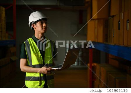Male manager in hardhat and vest using laptop, checking inventory production stock control in warehouse 105902156