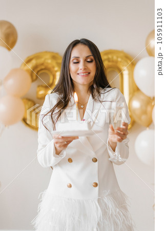 Young caucasian woman holding a cake with a candle and a glass of champagne in honor of the thirtieth birthday in a stylish white dress with feathers 105911103