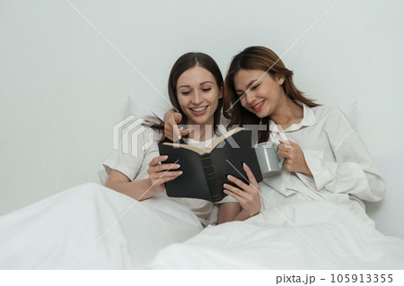 Two Asian cheerful happy female girlfriends having a coffee sitting on the bed while looking at the book. Concept lifestyle lesbian couple Two Asian cheerful happy female girlfriends having a coffee sitting on the bed while looking at the book. Concept lifestyle lesbian couple 105913355