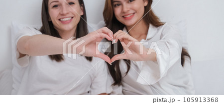 Lesbian couple together concept. Portrait of a smiling young lesbian couple other while sitting together and hand made heart symbol in the morning at bedroom 105913368