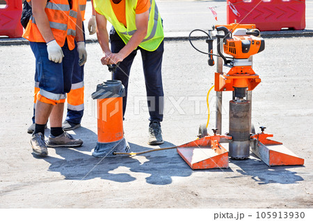 Road workers in reflective raincoats drill asphalt with a core drill. 105913930