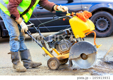 A road worker in reflective overalls uses a portable asphalt cutter to cut away worn asphalt with a diamond blade. 105913931