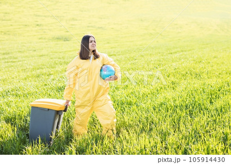 A woman in a yellow protective suit stands in the middle of a green field holding a globe, and with her other hand carries a garbage can. The concept of saving the planet from environmental pollution 105914430