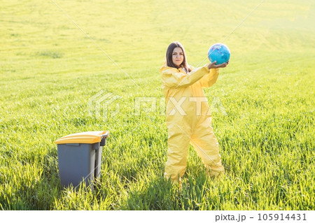 A woman in a yellow protective suit stands in the middle of a green field and holds a globe in her hands, next to a garbage can A woman in a yellow protective suit stands in the middle of a green field and holds a globe in her hands, next to a garbage can 105914431