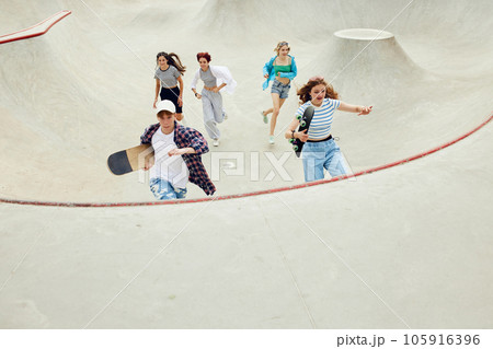 Group of teens in casual clothes, boy and girls running with skate on skateboard ramp. Activity and fun 105916396