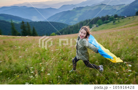 Cute girl running in mountains with Ukrainian flag. Little girl smiling at camera with Ukrainian flag on her shoulders. Feeling freedom in young people. 105916428