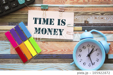 On a wooden background an alarm clock, stickers and a white sheet with the inscription time is money 105922872