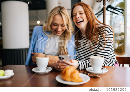 Portrait of two cheerful beautiful women friends sitting in cafe indoors, looking at phone and talking, laughing. Happy pretty females girlfriends using social media on smartphone in coffee shop. Portrait of two cheerful beautiful women friends sitting in cafe indoors, looking at phone and talking, laughing. Happy pretty females girlfriends using social media on smartphone in coffee shop. 105923292