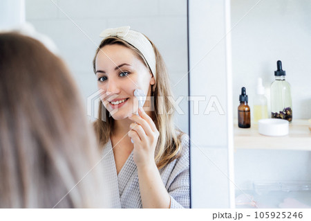 Young woman in bathrobe looking in mirror and making face massage with micro needle meso derma Roller. concept of natural procedures for skin care. Home Beauty self-care treatment. Selective focus. 105925246