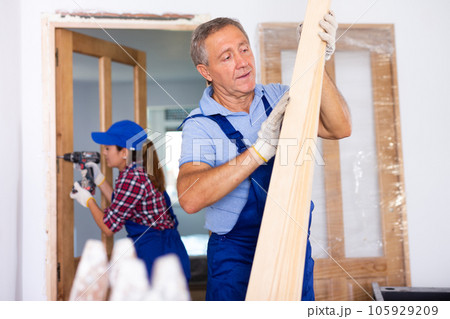 Middle-aged male carpenter in a blue uniform checking wooden planks indoors 105929209