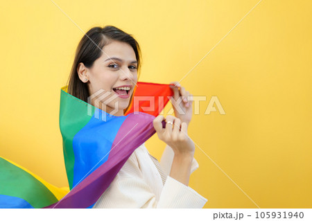 Happy cheerful caucasian white woman smiling and holding a rainbow flag which is the symbolic of LGBTQ people on yellow background. The diversity in genders and lifestyles concept. 105931940