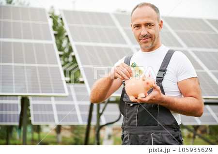Investor putting euros to piggy bank on background of solar panels. Knowledgeable man investing in green energy for future profit. Male adult in overalls happy about investment he did. Investor putting euros to piggy bank on background of solar panels. Knowledgeable man investing in green energy for future profit. Male adult in overalls happy about investment he did. 105935629