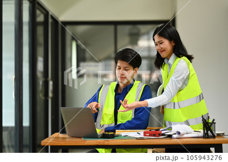 Two engineers in reflective vests working on blueprint and discussing the new project in office. 105943276