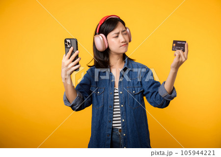 Portrait young asian woman with doubt face wearing denim clothes and headphone holding smartphone and credit card. Shopping online mobile phone entertainment lifestyle concept. Portrait young asian woman with doubt face wearing denim clothes and headphone holding smartphone and credit card. Shopping online mobile phone entertainment lifestyle concept. 105944221