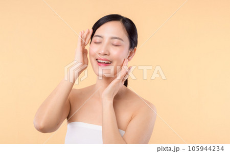 Beautiful girl asian model touching fresh glowing hydrated facial skin on beige background closeup. Beauty face young woman with natural makeup and healthy skin portrait. Skin care concept 105944234