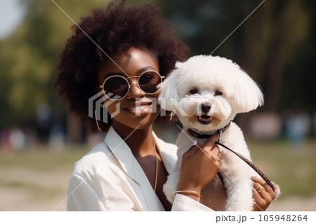 Happy young African American woman is holding her Bichon Frise dog on her hands in the street. AI generated. 105948264
