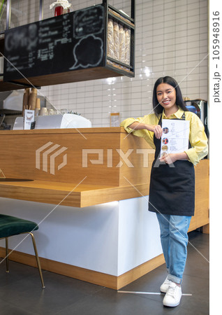 Cute smiling waitress in a black apron standing near the cafe counter 105948916