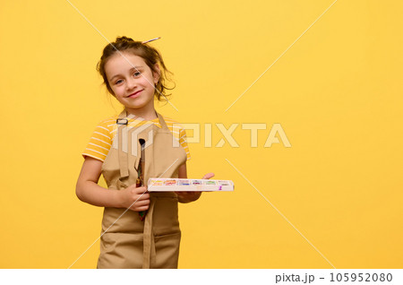 Adorable baby girl in beige apron, talented painter posing with paintbrush and watercolors smiling looking at camera Adorable baby girl in beige apron, talented painter posing with paintbrush and watercolors smiling looking at camera 105952080