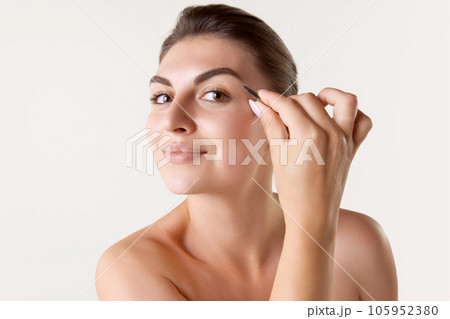 Beautiful young woman plucking eyebrows with tweezers against white studio background Beautiful young woman plucking eyebrows with tweezers against white studio background 105952380