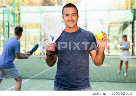Portrait of a satisfied adult man with racket and ball in his hands on padel court 105962019