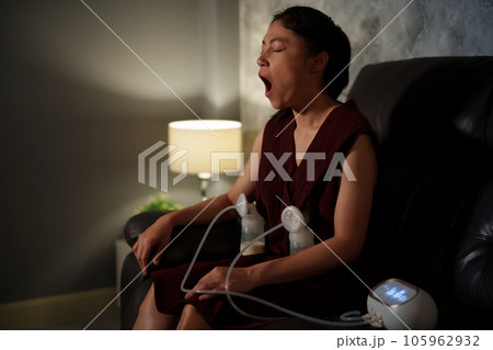 sleepy mother yawning and using breast pump machine to pumping milk for baby on sofa in the living room at night 105962932