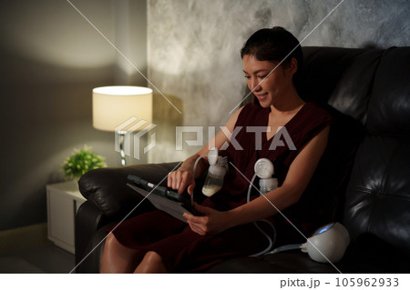 mother using tablet and breast pump machine to pumping milk for baby on sofa in the living room at night 105962933