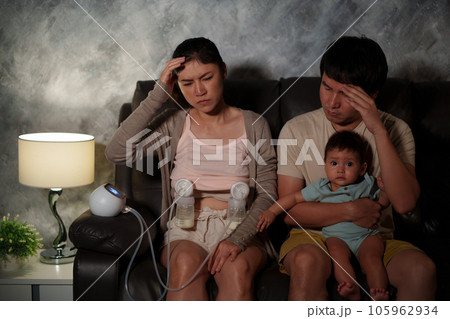 stressed father and mother using breast pump machine to pumping milk for infant baby on sofa in the living room at night 105962934