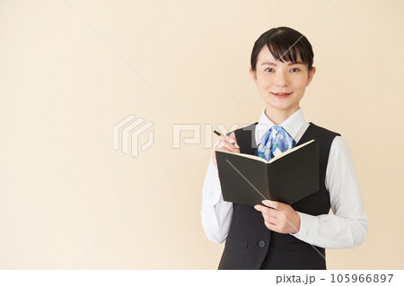 手帳を書くベストにリボンタイの制服を着た、受付業務、事務業務の女性 手帳を書くベストにリボンタイの制服を着た、受付業務、事務業務の女性 105966897