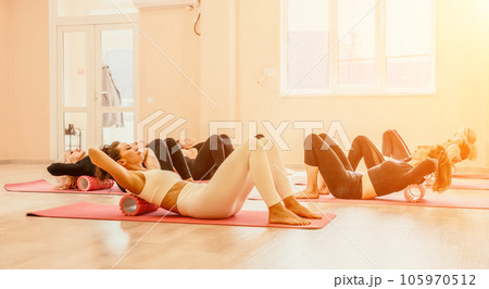 Happy middle aged well looking women, performing fascia exercises on floor using massage foam roller - tool to relieve tension in the back and relieve muscle pain. Female fitness yoga routine concept 105970512