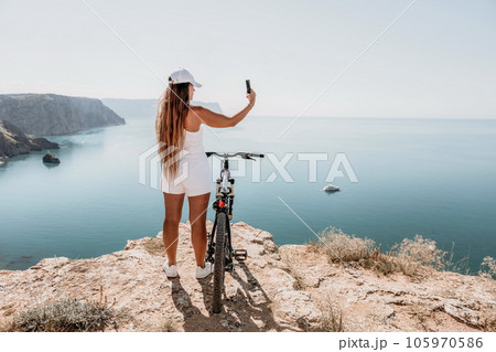 A woman cyclist on a mountain bike looking at the landscape of mountains and sea. Rear view of cyclist woman standing in front to the sea with outstretched arms. Freedom and healthy lifestyle concept. A woman cyclist on a mountain bike looking at the landscape of mountains and sea. Rear view of cyclist woman standing in front to the sea with outstretched arms. Freedom and healthy lifestyle concept. 105970586