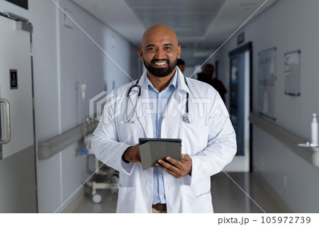Portrait of happy biracial male doctor wearing lab coat holding tablet in corridor at hospital Portrait of happy biracial male doctor wearing lab coat holding tablet in corridor at hospital 105972739