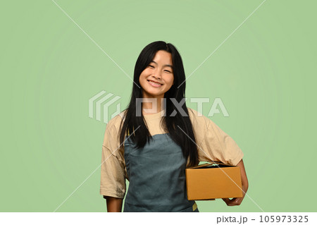 Cute teenage Asian girl holding parcel box isolated on green background. Post, package, shipping and delivery concept. Cute teenage Asian girl holding parcel box isolated on green background. Post, package, shipping and delivery concept. 105973325
