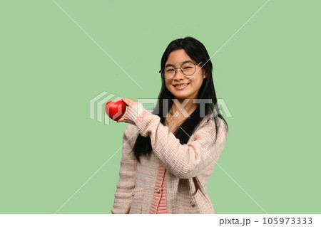 Smiling Asian teenage girl holding red heart isolated on green background. Love, valentine, gift and health symbol. Smiling Asian teenage girl holding red heart isolated on green background. Love, valentine, gift and health symbol. 105973333