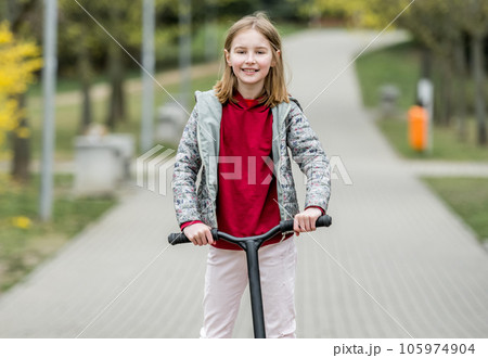 Little cute girl riding a scooter on a path in the park, smiling and looking into the camera 105974904