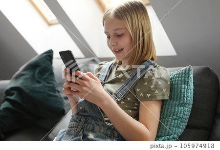 Child girl with smartphone sitting on sofa at home and chatting with friend in social media. Pretty preteen kid with mobile phone texting Child girl with smartphone sitting on sofa at home and chatting with friend in social media. Pretty preteen kid with mobile phone texting 105974918