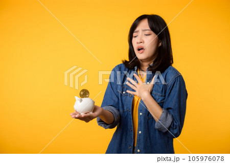 Unhappy asian young woman wearing yellow t-shirt denim shirt pulling digital coin crypto currency out of piggy bank isolated on yellow background. Payment digital money debt financial concept. Unhappy asian young woman wearing yellow t-shirt denim shirt pulling digital coin crypto currency out of piggy bank isolated on yellow background. Payment digital money debt financial concept. 105976078