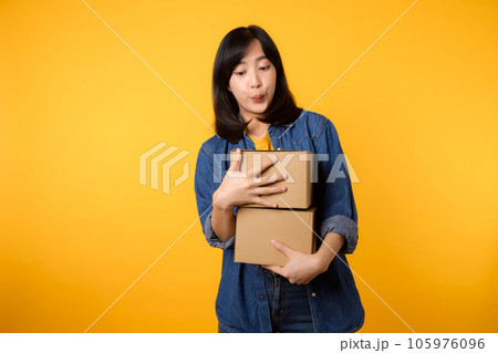 Portrait asian young woman wearing yellow t-shirt and denim shirt holding parcel box isolated on yellow studio background, Delivery courier and shipping service concept. Portrait asian young woman wearing yellow t-shirt and denim shirt holding parcel box isolated on yellow studio background, Delivery courier and shipping service concept. 105976096