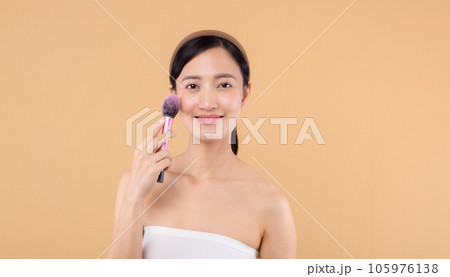 Portrait happy young asian woman with natural make up face holding cosmetic skin powder blusher isolated on beige background. Female apply skincare brush treatment. beauty product cosmetology concept. Portrait happy young asian woman with natural make up face holding cosmetic skin powder blusher isolated on beige background. Female apply skincare brush treatment. beauty product cosmetology concept. 105976138