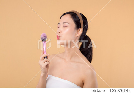 Portrait happy young asian woman with natural make up face holding cosmetic skin powder blusher isolated on beige background. Female apply skincare brush treatment. beauty product cosmetology concept. Portrait happy young asian woman with natural make up face holding cosmetic skin powder blusher isolated on beige background. Female apply skincare brush treatment. beauty product cosmetology concept. 105976142