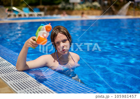 Young pretty caucasian girl with blue eyes shoots with a water gun in the pool. Pretty girl playing with water guns in pool. Cheerful children playing waterguns, rejoicing, jumping, swimming in pool. Young pretty caucasian girl with blue eyes shoots with a water gun in the pool. Pretty girl playing with water guns in pool. Cheerful children playing waterguns, rejoicing, jumping, swimming in pool. 105981457