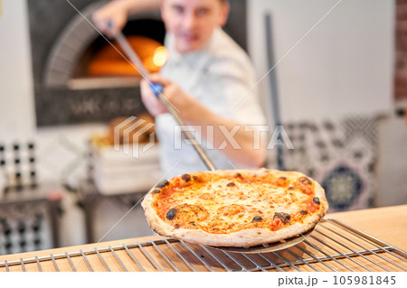 The chef prepares pizza. Raw pizza ready to bake. Cook in a blue apron in the kitchen. with a shovel in his hands. boxes for food delivery on background. The chef prepares pizza. Raw pizza ready to bake. Cook in a blue apron in the kitchen. with a shovel in his hands. boxes for food delivery on background. 105981845