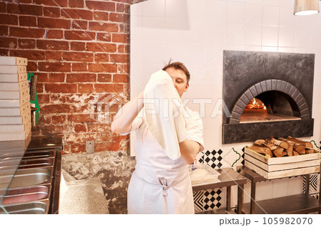 The chef prepare pizza. Raw pizza ready to bake. Cook in a apron in the kitchen Rolls out pizza dough. boxes for food delivery on background. The chef prepare pizza. Raw pizza ready to bake. Cook in a apron in the kitchen Rolls out pizza dough. boxes for food delivery on background. 105982070