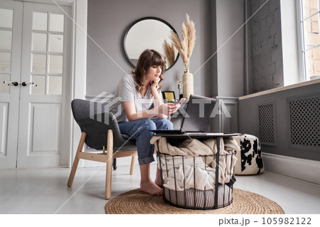 Home office. Beautiful cheerful girl using black laptop while sitting on chair in living room at home. Young casual woman using laptop for communication and work.  105982122