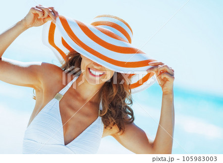 smiling woman in swimsuit hiding behind beach hat 105983003