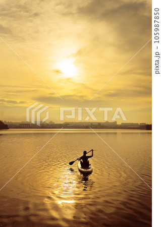 Stand up kayaking or stand-up paddleboarding on a quiet lake at sunset during a warm summer beach holiday, active woman. Stand up kayaking or stand-up paddleboarding on a quiet lake at sunset during a warm summer beach holiday, active woman. 105987850