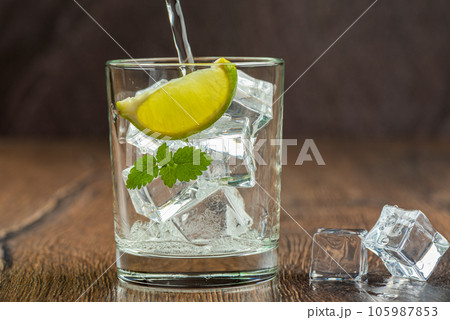 Refreshing water is poured into a glass with ice cubes, lime wedge and mint leaves. 105987853