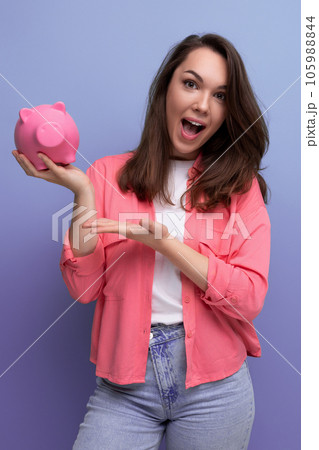 positive brunette young woman in a shirt and jeans holding a piggy bank with savings positive brunette young woman in a shirt and jeans holding a piggy bank with savings 105988844