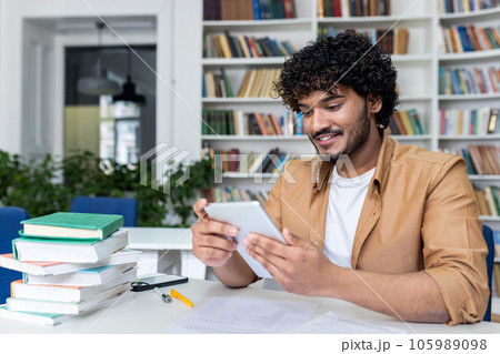 Young successful student studying sitting inside university academic library, man smiling holding tablet computer, Hispanic man using study app. 105989098