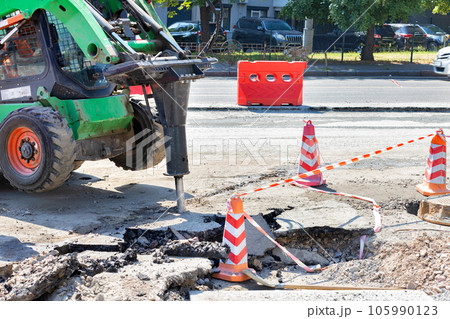 The barrel of a powerful jackhammer on a maneuverable and nimble road tractor breaks the old asphalt on a summer day. 105990123